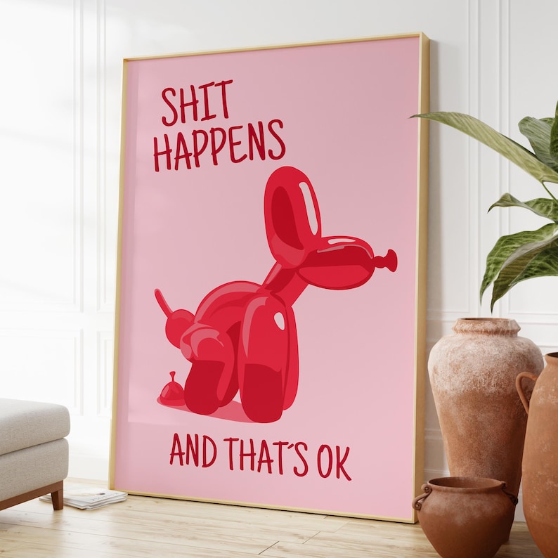 May include: A framed print with the text "SHIT HAPPENS AND THAT'S OK" on a pink background. A red balloon dog is featured in the center. The print is in a gold frame and is displayed in a room with neutral tones.