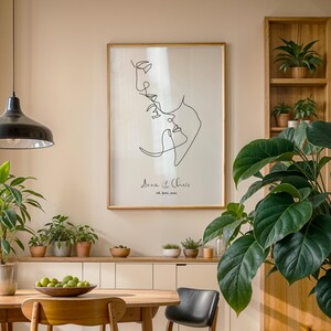 May include: A black and white line drawing of a couple's faces, with the text "Ava & Chris 08. June. 2021" below the drawing. The drawing is framed and hanging on a wall in a room with a wooden table and chairs, plants, and a shelf with plants.