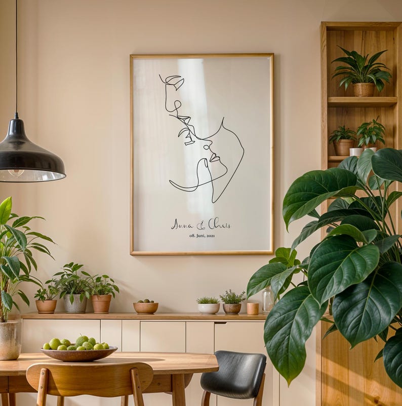 May include: A black and white line drawing of a couple's faces, with the text "Ava & Chris 08. June. 2021" below the drawing. The drawing is framed and hanging on a wall in a room with a wooden table and chairs, plants, and a shelf with plants.