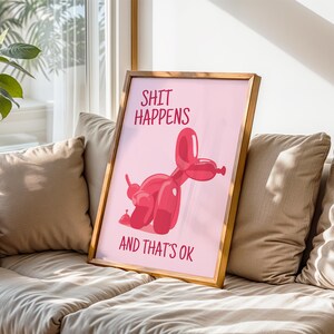 May include: A framed print with a pink background and a red balloon dog illustration. The text "SHIT HAPPENS" is at the top, and "AND THAT'S OK" is at the bottom. The frame is a light brown color.