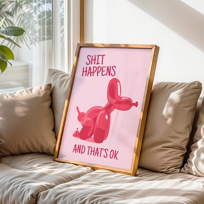 May include: A framed print with a pink background and a red balloon dog illustration. The text "SHIT HAPPENS" is at the top, and "AND THAT'S OK" is at the bottom. The frame is a light brown color.