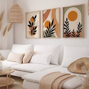 Puede incluir: Tres impresiones de arte abstracto de estilo minimalista con siluetas negras de hojas y ramas sobre un fondo de colores naranja, marr&oacute;n y verde apagados. Las impresiones est&aacute;n colgadas en una pared blanca sobre un sof&aacute; blanco con almohadas y una manta.