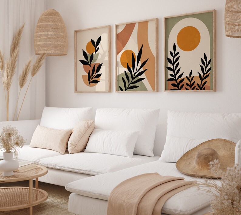 Puede incluir: Tres impresiones de arte abstracto de estilo minimalista con siluetas negras de hojas y ramas sobre un fondo de colores naranja, marr&oacute;n y verde apagados. Las impresiones est&aacute;n colgadas en una pared blanca sobre un sof&aacute; blanco con almohadas y una manta.