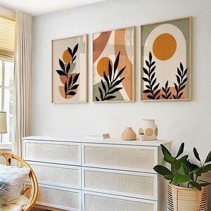 Puede incluir: Tres impresiones de arte abstracto con un dise&ntilde;o minimalista que presenta hojas negras, c&iacute;rculos naranja y marr&oacute;n y un fondo blanco. Las impresiones est&aacute;n enmarcadas en madera natural y colgadas en una pared blanca sobre una c&oacute;moda blanca con cajones de mimbre.