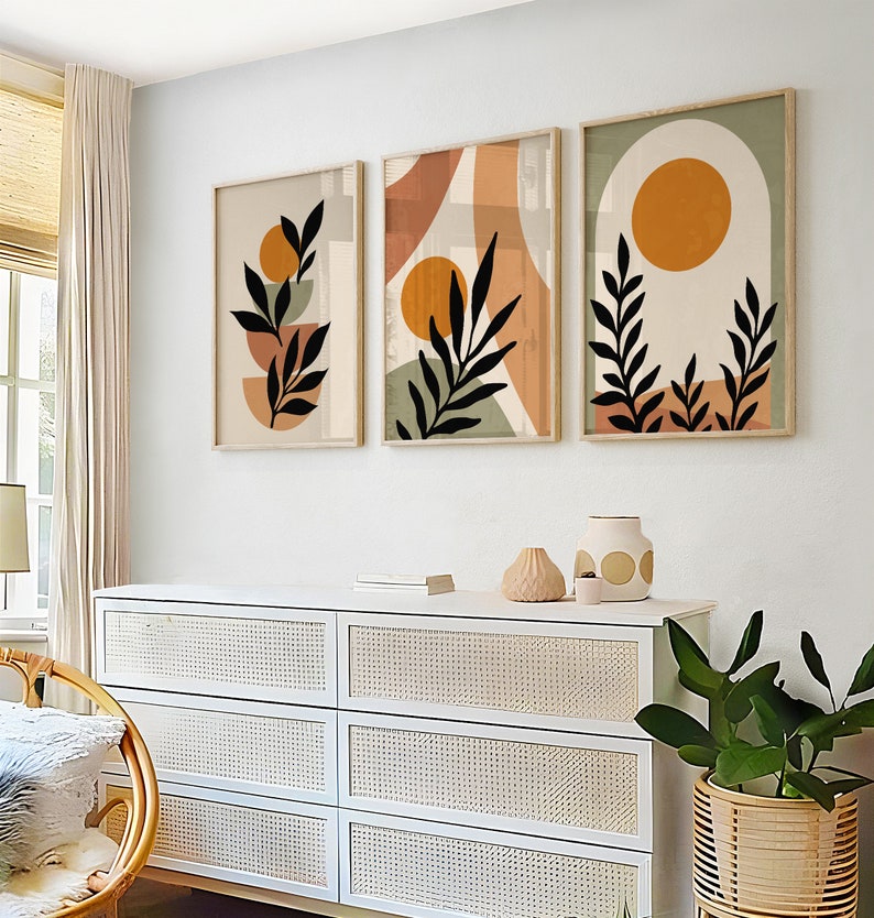Puede incluir: Tres impresiones de arte abstracto con un dise&ntilde;o minimalista que presenta hojas negras, c&iacute;rculos naranja y marr&oacute;n y un fondo blanco. Las impresiones est&aacute;n enmarcadas en madera natural y colgadas en una pared blanca sobre una c&oacute;moda blanca con cajones de mimbre.