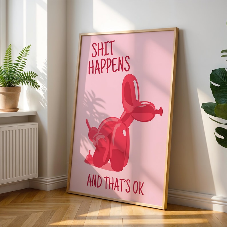 May include: A framed pink poster with the words "SHIT HAPPENS AND THAT'S OK" in red. A red balloon dog is featured on the poster. The poster is leaning against a wall next to a window with a potted plant.