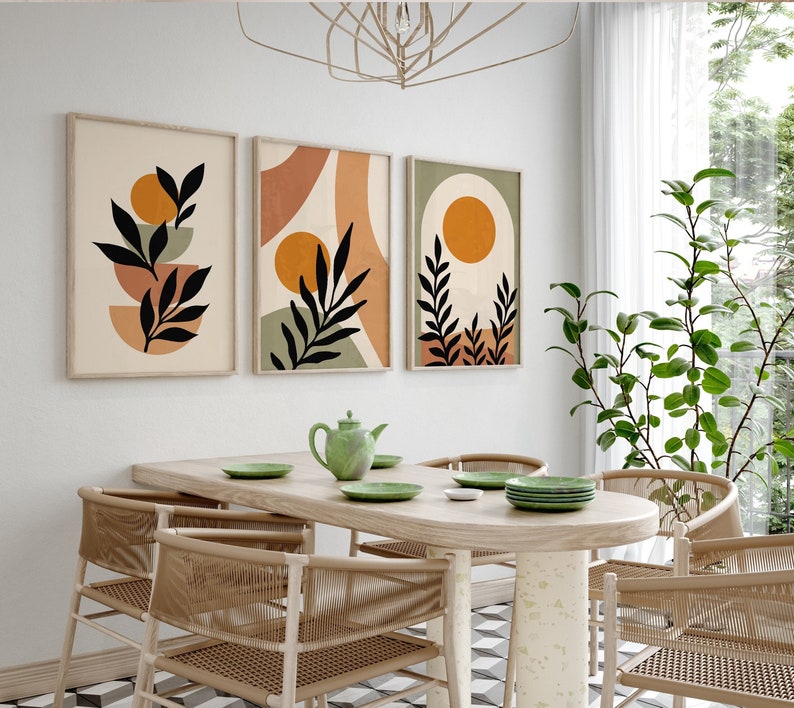 Puede incluir: Tres impresiones de arte abstracto con un dise&ntilde;o minimalista que presenta hojas negras, c&iacute;rculos naranjas y formas geom&eacute;tricas en tonos marrones, naranjas y verdes. Las impresiones est&aacute;n enmarcadas y colgadas en una pared blanca sobre una mesa de comedor de madera con cuatro sillas.