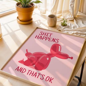 May include: A framed print with the text "SHIT HAPPENS AND THAT'S OK" in red. The print features a red balloon animal on a pink background. The frame is made of light-colored wood.