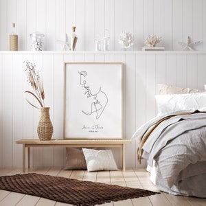 May include: A black and white line drawing of a couple kissing, with the text "Anna & Chris" and the date "01. June. 2020" below the drawing. The drawing is framed and hanging on a white wall.