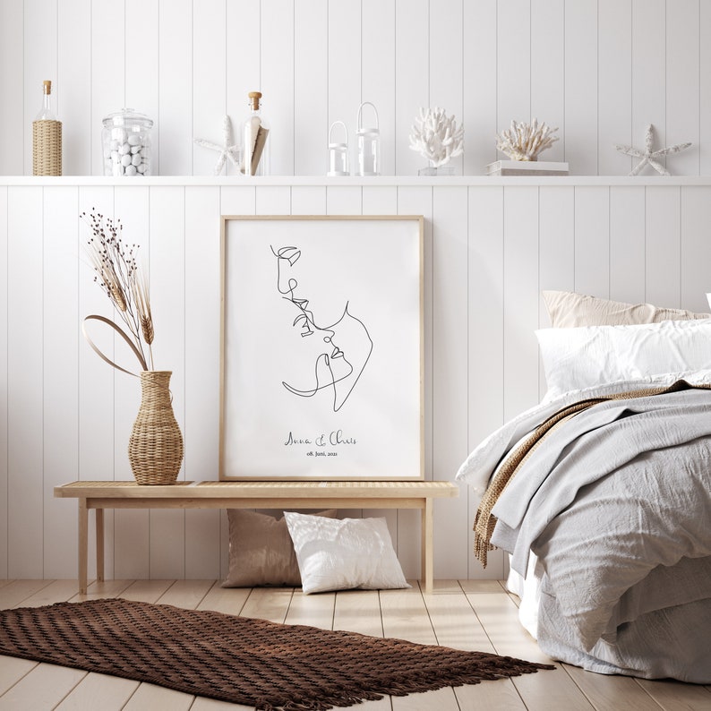 May include: A black and white line drawing of a couple kissing, with the text "Anna & Chris" and the date "01. June. 2020" below the drawing. The drawing is framed and hanging on a white wall.