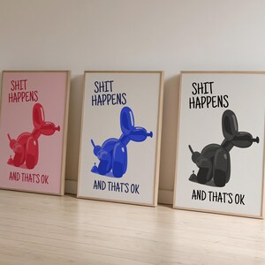 May include: Three framed art prints with the text "SHIT HAPPENS AND THAT'S OK". Each print features a balloon dog in red, blue, and black. The prints have a light wood frame and are leaning against a white wall.