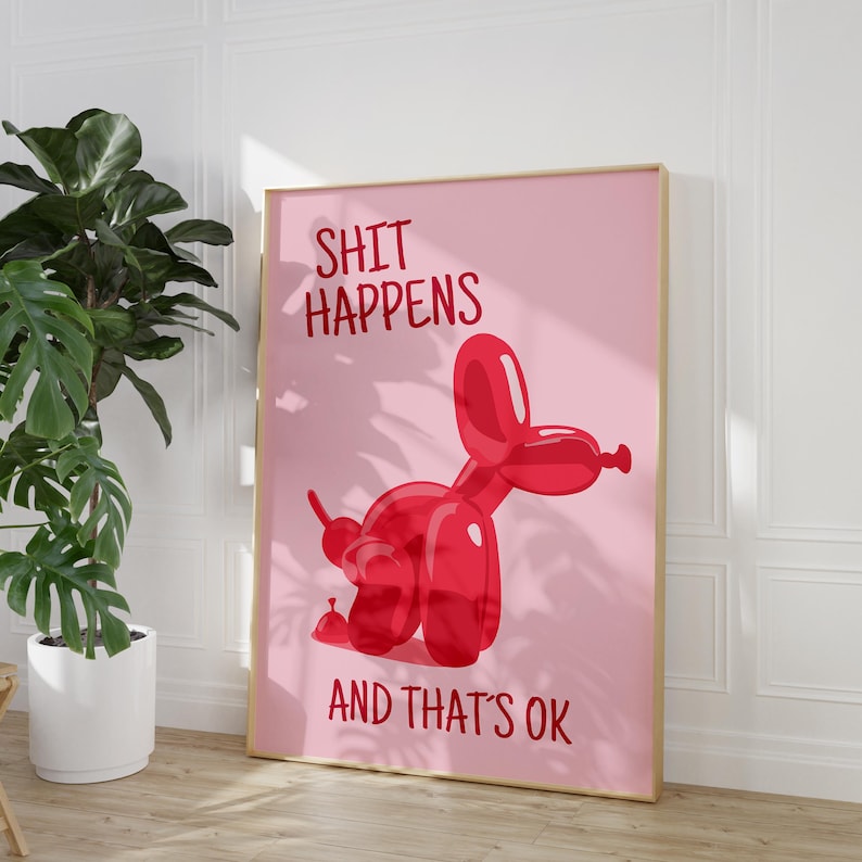 May include: A framed pink art print featuring a red balloon dog illustration. The text "SHIT HAPPENS AND THAT'S OK" is displayed in red. The artwork is displayed on a light wood floor.