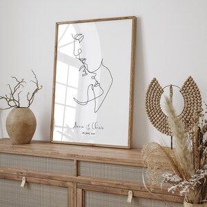 May include: A black and white line drawing of a couple's faces, with the text "Anna & Chris 08. June. 2021" below the drawing. The drawing is framed in a light wood frame and is hanging on a wall.
