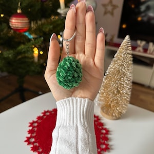 Könnte beinhalten: Ein grüner, plüschiger Weihnachtsbaum-Schlüsselanhänger. Der Schlüsselanhänger wird vor einem Weihnachtsbaum mit Ornamenten und Lichtern gehalten. Im Hintergrund befindet sich ein kleiner, goldfarbener Deko-Baum.