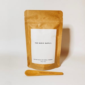 May include: A brown paper pouch with a white label that reads "THE BASIC SUPPLY." and "HEATED SCALLOP SHELL POWDER 100g/3.5oz". A wooden spoon with the text "THE BASIC SUPPLY." printed on it is in front of the pouch.