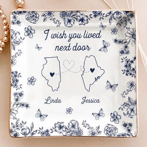 May include: A white ceramic square trinket dish with a gold rim, featuring a blue floral design and the text "I wish you lived next door." The dish includes outlines of two states, connected by a dotted line of hearts, with the names "Linda" and "Jessica" below.