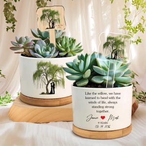 Może przedstawiać: Dwie białe ceramiczne donice z sukulentami, z motywem wierzby płaczącej i napisem "Jak wierzba, nauczyliśmy się zginać z wiatrami życia, zawsze mocno razem." Zawiera drewniane paliki.
