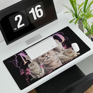 May include: A black rectangular desk mat featuring a design of several gray cats with yellow eyes. The word "MO" is in pink and gold, with pink lightning bolts. A white keyboard and mouse are on top.
