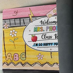 May include: A yellow and pink welcome mat with the text "Welcome to Mrs. Phoe's Class K. I'm so happy you're here!"  The mat has a pencil, ruler, apple, and ABCs on it.