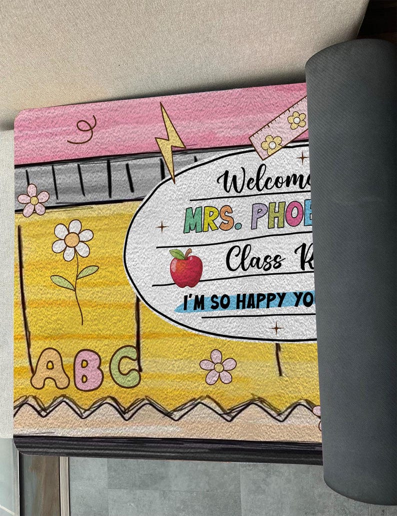 May include: A yellow and pink welcome mat with the text "Welcome to Mrs. Phoe's Class K. I'm so happy you're here!"  The mat has a pencil, ruler, apple, and ABCs on it.