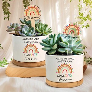 Op de afbeelding: Twee witte keramische plantenpotten met vetplanten, elk met een regenboogontwerp en de tekst "MAKING THE WORLD A BETTER PLACE SINCE 1975 Jessica." Houten en heldere acryl plantenstokken met de naam "Jessica" zijn ingevoegd.