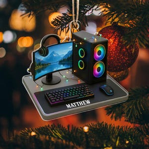 May include: A gaming setup Christmas ornament featuring a curved monitor, keyboard, mouse, and PC tower. The keyboard has the name "MATTHEW" on it. The PC tower and keyboard have colorful LED lights. A headset is placed on the monitor.