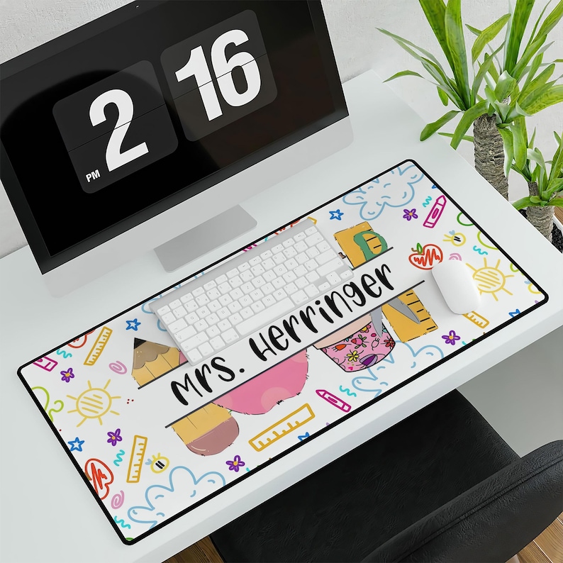 May include: A white desk mat with a colorful design featuring a pencil, a ruler, and a rainbow. The text "mrs. Herringer" is written in black letters on the mat.