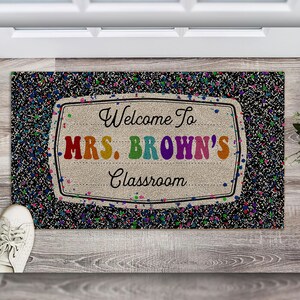 May include: Welcome to Mrs. Brown's Classroom doormat with a black background and colorful confetti. The text is in a white frame with a burlap texture.
