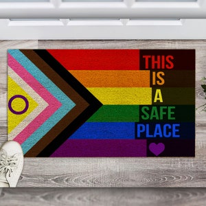 May include: A doormat with a rainbow flag design and the text "This is a safe place" with a purple heart. The flag includes the colors red, orange, yellow, green, blue, indigo, and violet.