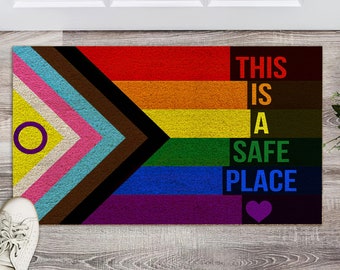 May include: A doormat with a rainbow flag design and the text "This is a safe place" with a purple heart. The flag includes the colors red, orange, yellow, green, blue, indigo, and violet.