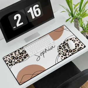 May include: A white desk mat with a leopard print design, abstract lines, and two brown circles. The text "Sophia" is written in black cursive on the mat. A white keyboard and a computer mouse are on the mat.
