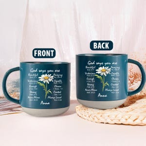 May include: Two teal ceramic mugs with a white and yellow daisy design. The mugs have the text "God says you are" and inspirational words. The mugs have a gray base and a dark teal handle. The name "Anna" is printed on the mugs.