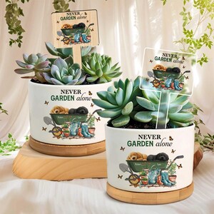 Może przedstawiać: Dwie białe ceramiczne donice z sukulentami, każda z napisem "NEVER GARDEN alone". Donice mają wzór szczeniąt w taczce z narzędziami ogrodniczymi. Drewniane znaki z tym samym tekstem są umieszczone w donicach.