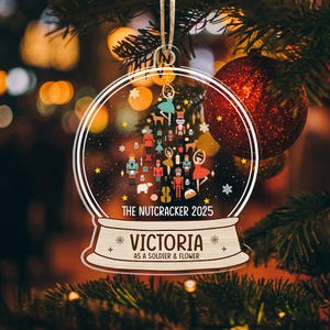 Pode incluir: Enfeite de Natal em acrílico transparente com design de globo de neve. O enfeite apresenta um tema Quebra-Nozes com o texto "THE NUTCRACKER 2025" e "VICTORIA AS A SOLDIER & FLOWER". O enfeite está pendurado em uma árvore.