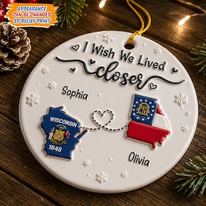 Op de afbeelding: Een rond wit keramieken ornament met de tekst I Wish We Lived closer, de namen Sophia en Olivia, en twee 3D-reliëfkaarten van Wisconsin en Georgia verbonden door een stippellijn in hartvorm, versierd met kleine sneeuwvlokken en een gouden koord.