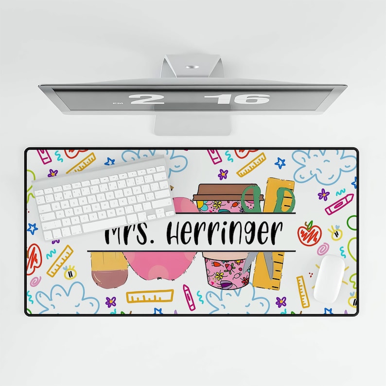 May include: White desk mat with a colorful doodle design featuring a pencil, apple, and coffee cup. The text "Mrs. Herringer" is written in a black banner across the middle of the mat.