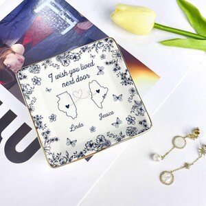 May include: Square ceramic trinket dish with a floral and butterfly design. The dish has a gold rim and features the text "I wish you lived next door" with two state outlines and names. A decorative item for a desk or dresser.
