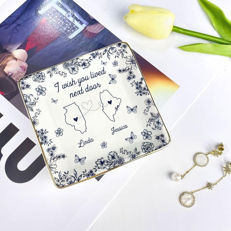 May include: Square ceramic trinket dish with a floral and butterfly design. The dish has a gold rim and features the text "I wish you lived next door" with two state outlines and names. A decorative item for a desk or dresser.
