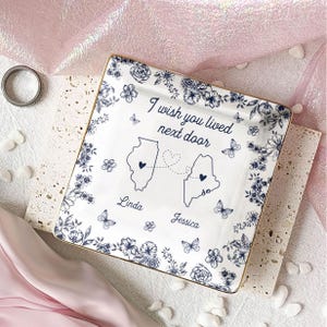May include: Square white ceramic dish with a blue floral design and gold trim. The dish features the text "I wish you lived next door" and outlines of two states connected by a dotted line. The names "Linda" and "Jessica" are below the states.