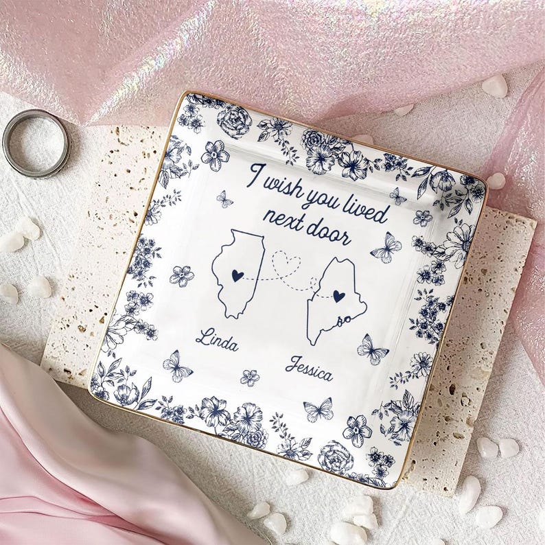 May include: Square white ceramic dish with a blue floral design and gold trim. The dish features the text "I wish you lived next door" and outlines of two states connected by a dotted line. The names "Linda" and "Jessica" are below the states.
