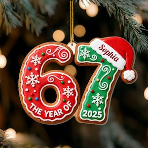 May include: A festive Christmas ornament shaped like the numbers 67, designed to resemble gingerbread cookies. The number 6 is red with white icing and snowflakes, while the 7 is green with a Santa hat and the name "Sophia". The year 2025 is also displayed.