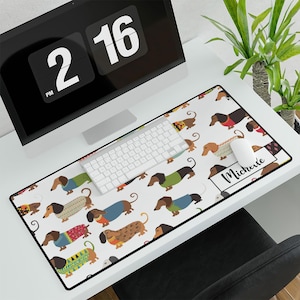 May include: A white desk mat with a repeating pattern of dachshund dogs wearing sweaters. The dogs are in various colors and patterns. The mat has a personalized name, "Michelle", in a white box with a black border.