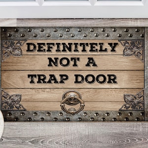 May include: A brown wooden doormat with a metal frame and rivets. The doormat has the text "DEFINITELY NOT A TRAP DOOR" in black lettering.