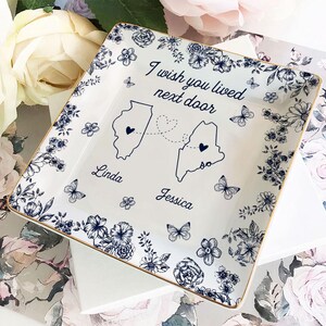 May include: Square white ceramic dish with a gold rim, featuring a floral and butterfly design in navy blue. The dish has the text "I wish you lived next door" with two state outlines and names.