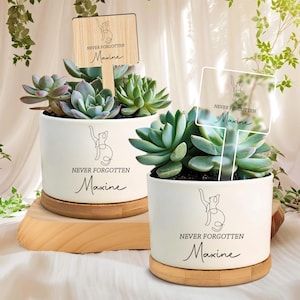 Könnte beinhalten: Zwei weiße Keramik-Pflanztöpfe mit Sukkulenten, jeweils auf einem Holzsockel. Die Töpfe tragen den Text "NEVER FORGOTTEN Maxine" und eine Katzenillustration. Ein Topf hat ein Holzschild, der andere ein klares Schild.