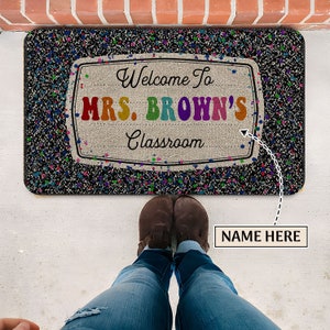 May include: Welcome to Mrs. Brown's Classroom doormat with a black background and colorful confetti. The doormat has a white rectangle with the text "Welcome to Mrs. Brown's Classroom" in rainbow colors.