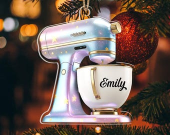 May include: A Christmas tree ornament shaped like a stand mixer. The ornament is a pastel blue and purple with gold accents and star details. The white bowl has the name "Emily" in black script.