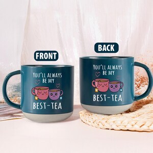 Może przedstawiać: Turkusowy ceramiczny kubek z szarą podstawą, z napisem "YOU'LL ALWAYS BE MY BEST-TEA" i grafiką dwóch uśmiechniętych filiżanek. Kubek ma duży uchwyt i jest przeznaczony do gorących napojów.