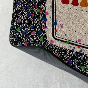 May include: A black and white patterned doormat with colorful paint splatters. The doormat has a black border and a white background with the word "Make" in rainbow letters.