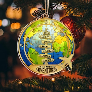 Op de afbeelding: Een globe-vormige kerstversiering met een gouden rand en een wereldkaartontwerp. De versiering heeft pijlen die naar verschillende landen wijzen, het jaar 2025 en de tekst "ANNIE & MATTHEW ADVENTURES". Een gouden vliegtuigafbeelding is ook inbegrepen.
